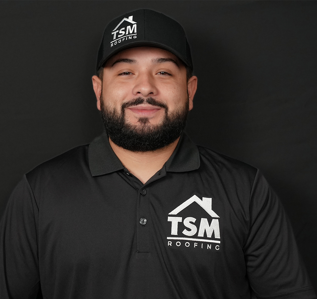 Smiling man wearing TSM Roofing logo shirt and cap against dark background.