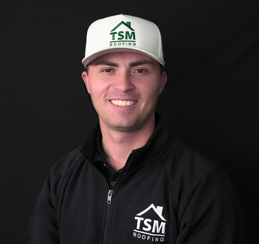 Smiling man wearing a TSM Roofing hat and jacket against dark background.