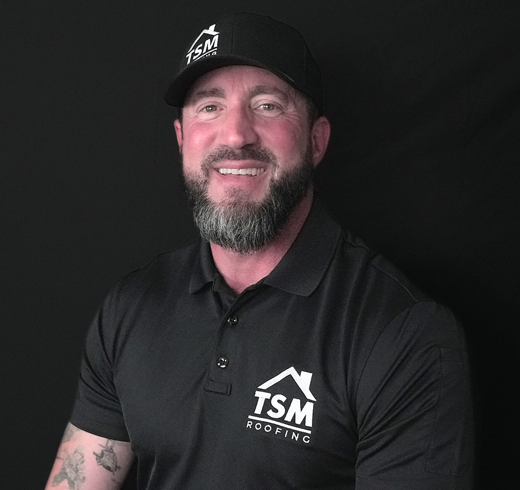 Smiling man wearing TSM Roofing shirt and cap on black background.