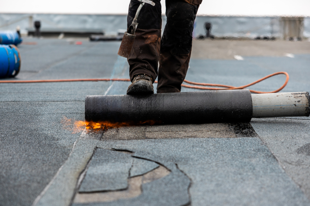 Perofessional roofer at work securing rolled tar paper with a blowtorch on a flat roof repair process.